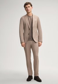 Beige Leinenanzug mit tailored Fit, einreihiger Jacke und Reverskragen; kombiniert mit einem schokoladenfarbigen Strickhemd und dunklen Stiefeln.