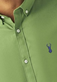 Grünes Hemd mit Kragen aus Baumwolle, ausgestattet mit weißen Knöpfen und einem kleinen blauen gestickten Hirschlogo auf der Brust. Glatte Textur.