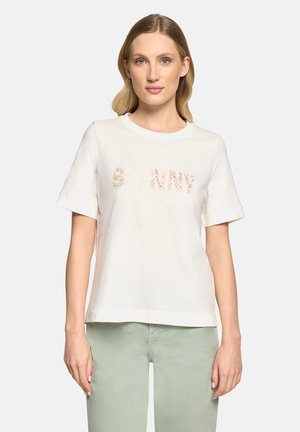 Lyshåret kvinde iført en hvid kortærmet T-shirt med let mønstret tekst og lysegrønne bukser, stående mod en ensfarvet baggrund.