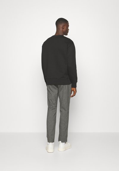 Zwarte sweatshirt met een ronde hals, gecombineerd met grijze broeken. Model draagt witte sneakers. De focus ligt op strakke lijnen en een minimalistisch ontwerp.