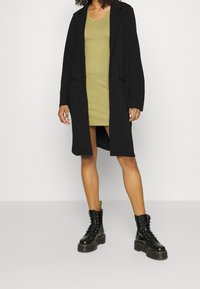 Czarny długi płaszcz z otwartym przodem, dwoma kieszeniami, zestawiony z zieloną sukienką i czarnymi sznurowanymi butami wojskowymi z grubą podeszwą.