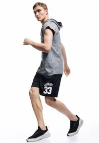 Grau ärmelloses Hoodie, schwarze Shorts mit dem Aufdruck "LEGENDARY 33" und schwarze Sportschuhe mit heller Sohle. Modell in Gehpose.