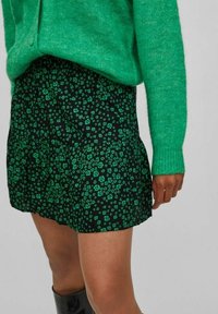 Pull vert en maille à manches longues associé à une jupe noire avec un motif floral vert et un design à volants.