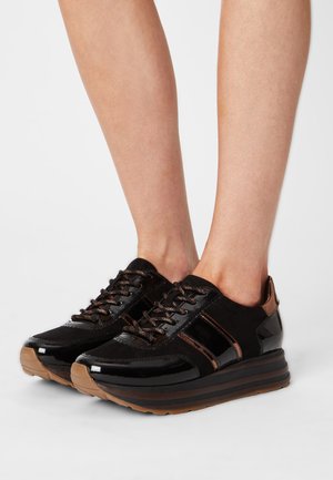 Nogi w czarnych trampkach na platformie z połyskującymi i matowymi wykończeniami, brązowymi akcentami i brązowymi podeszwami na białym tle.