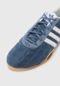 Sinine Adidas toss, millel on valged triibud, tekstureeritud õmblused, sametised detailid ja kummist tallad, vaadatuna ülalt-ees.