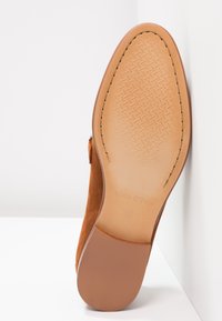 Semelle de chaussure en cuir marron avec motif antidérapant texturé, bords cousus et talon lisse, positionnée à la verticale sur fond blanc.