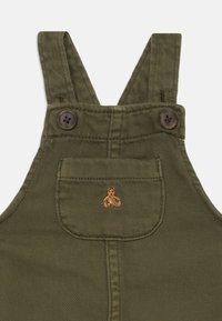 Olive zeleni bombažni kombinezon s širokimi naramnicami, rjavimi gumbi in žepom na prsih, ki ima majhen vezen bear dizajn.