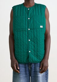 Chaleco sin mangas acolchado verde con dos bolsillos frontales y botones de presión plateados. Presenta un parche de logo en el lado izquierdo.