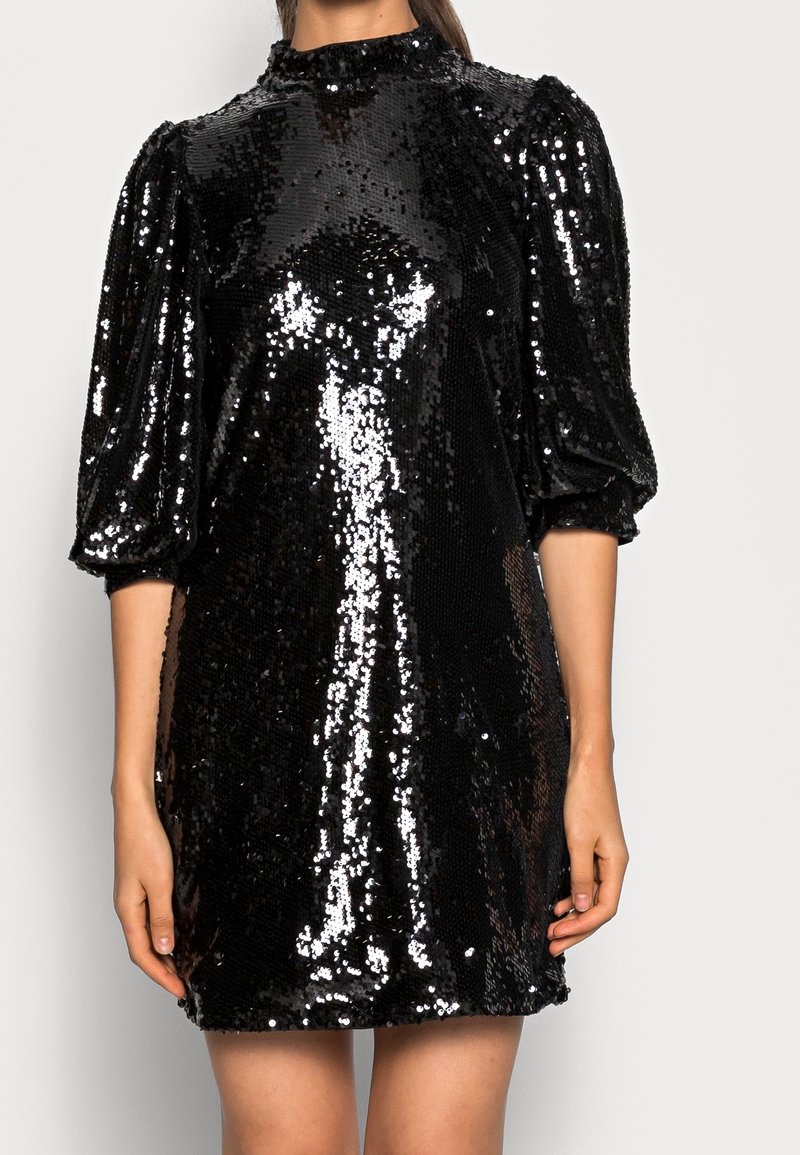 Vestido mini negro con lentejuelas, mangas abullonadas tres cuartos y cuello alto, llevado por una persona de pie sobre un fondo liso.