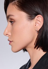 Srebrna kolczyk w kształcie obrączki o unikalnym wzorze noszony na lewej uchu. Krótkie, proste czarne włosy i gładka skóra widoczne w profilu.