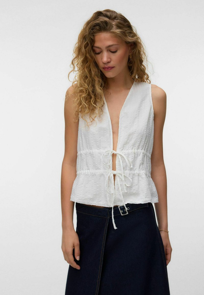 Femme aux cheveux blonds bouclés portant un haut blanc texturé sans manches avec des liens à l'avant et une jupe bleu foncé avec un détail boucle, regardant vers le bas.