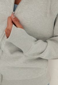 Lichtgrijze zip-up hoodie met een zachte textuur, ribgebreide manchetten en een zilveren rits. De stof lijkt glad en is geschikt voor casual kleding.