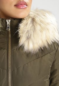 Chaqueta acolchada verde oliva con cierre delantero, que cuenta con un cuello de piel sintética en crema clara. Tejido texturizado con detalles de costura en forma de chevrón.
