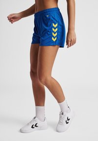 Blå idrottsshorts med ett gult chevronmönster på sidan. Elastisk midja, lätt material. Kombinerat med vita skor och strumpor.