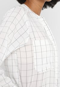 Bílá kostkovaná oversized košile s černým mřížkováním, kulatým výstřihem a kapsou na prsou, vyrobená z lehkého materiálu.