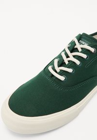 Grüne Segeltuch-Sneaker mit beiger Gummisohle, weißen Schnürsenkeln und kontrastierenden Ösen. Verfügt über ein strukturiertes Gewebe und gestickte Akzente.