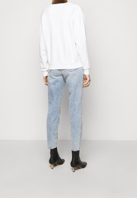 Personne portant un jean ajusté bleu clair, un haut à manches longues blanc et des bottines noires à talons, se tenant de dos.