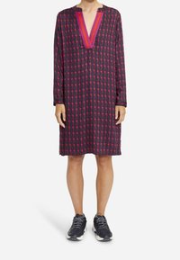 Sukienka z długim rękawem w ciemnym materiale z geometrycznym wzorem różowych i czerwonych kształtów. Posiada głęboki dekolt w serek oraz luźny krój. Noszona z trampkami.