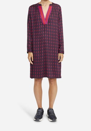 Robe à manches longues en tissu sombre avec un motif géométrique de formes roses et rouges. Elle présente un décolleté en V profond et une coupe décontractée. Portée avec des baskets.