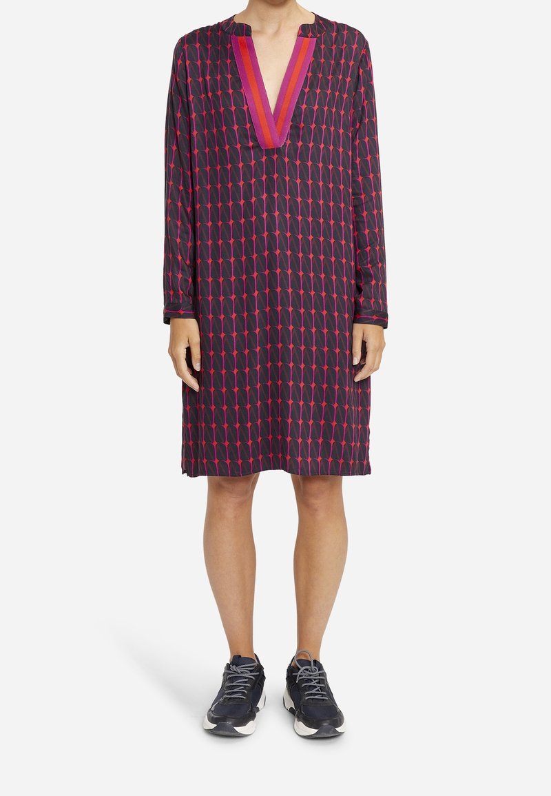 Sukienka z długim rękawem w ciemnym materiale z geometrycznym wzorem różowych i czerwonych kształtów. Posiada głęboki dekolt w serek oraz luźny krój. Noszona z trampkami.