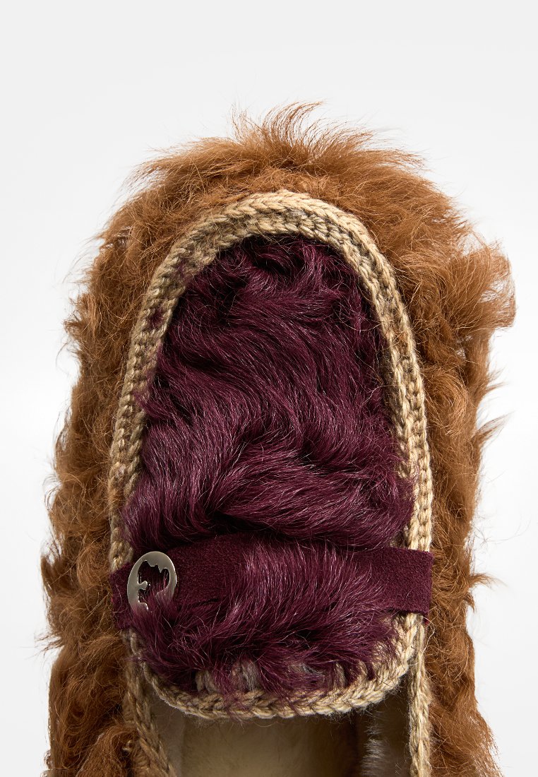 Braune, flauschige Hausschuhe mit maroonfarbenem Kunstfellinnenfutter, geflochtenem beige Trim und einem dekorativen silbernen Akzent auf einem dunklen Riemen.