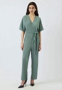 Jumpsuit plissé vert clair avec un design croisé et des manches larges, cintré à la taille avec une ceinture assortie. Associé à des talons noirs.
