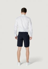Camicetta bianca a maniche lunghe, pantaloni corti blu navy e sneakers bianche. La camicia ha un col classico; i pantaloni corti sono sartoriali con tasche e un tessuto liscio.
