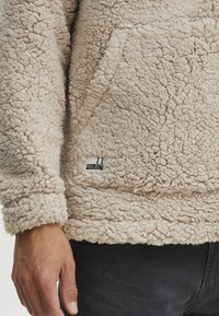 Beige fleece pullover with textured surface, kangaroo pocket, and small label featuring "11 PROJECT" in black text.