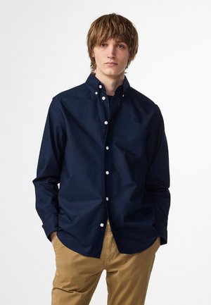 Marineblauwe button-up shirt met een kraag, lange mouwen en witte knopen. Stof lijkt glad en iets gestructureerd. Gecombineerd met kaki broek.