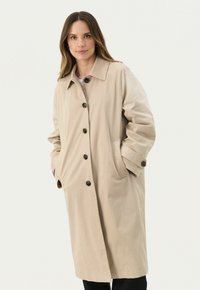 Beige színű trench coat, galléros nyakkal, öt gombos záródással, oldalsó zsebekkel és felgöngyölített ujjakkal, sötétbarna gombokkal.