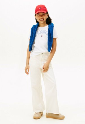 Blauwe kabelgebreide vest over een wit T-shirt met logo, roomkleurige wijde broek en tan schoenen. Rode pet met logo accentueert de outfit.