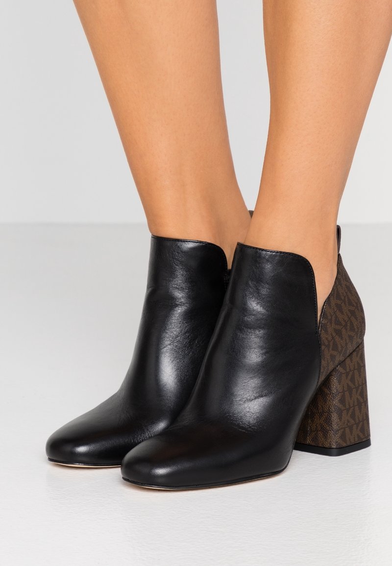 Bottines avec devant en cuir noir et talons carrés marron à motifs, portées sur des jambes nues devant un fond uni.