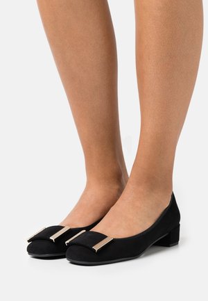 Zapatillas planas de ante negro con puntera redondeada, que presentan un detalle de acento en tono dorado en la parte delantera. Tacón bajo de bloque, textura suave, diseño sencillo.