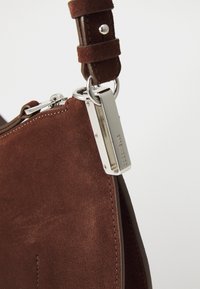 Borsa a mano in suede marrone con una tracolla in pelle liscia e minuteria in argento, dotata di chiusura zip e di un'etichetta metallica rettangolare.