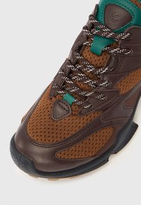 Brun sportssko med en kombination af glat og perforeret læder, kontrasterende turkis detaljer og en sort gummisål med tekstureret mønster.