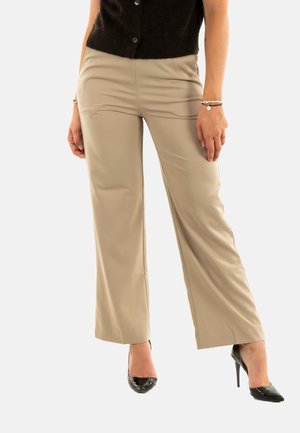 Femme portant un pantalon large beige, un haut sans manches noir, et des escarpins noirs à talons pointus, se tenant contre un fond uni.