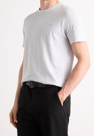 Mann trägt ein hellgraues T-Shirt mit Rundhalsausschnitt, das in schwarze Hose mit schwarzem Gürtel gesteckt ist, steht mit einer Hand in der Tasche.