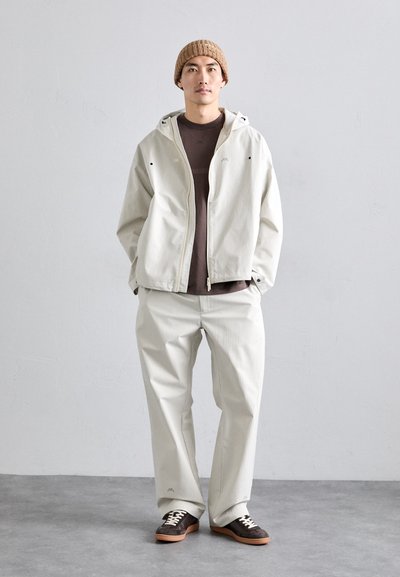 Lys beige hettejakke og matchende vide bukser sammen med en brun skjorte og brune joggesko. Strikket beige lue på hodet.