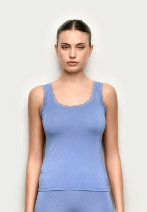 Młoda kobieta z zaczesanymi do tyłu włosami, ubrana w jasnoniebieską, bezrękawnikową bluzkę ozdobioną koronką, na jednolitym tle.