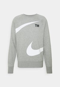 Grå sweatshirt med långa ärmar, med stora vita och svarta Nike-swoosh-designs och en liten "TM"-logotyp nära halsen. Mjukt material.