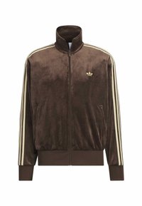 Velvet brown track jacket with high collar, full zipper, cream stripes on sleeves, and small gold logo on left chest. Two side pockets.