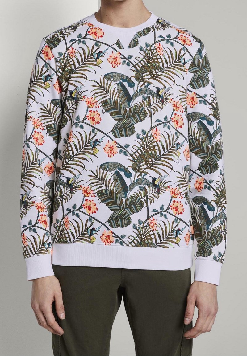 Sweat-shirt blanc à manches longues avec un motif floral tropical et des feuilles vertes, porté avec un pantalon vert olive.