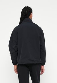 Persona con lunghe treccine che indossa un giubbotto imbottito nero e pantaloni neri, in piedi di spalle contro uno sfondo bianco uniforme.