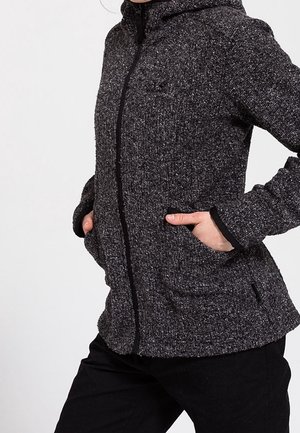 Osoba ubrana w ciemnoszarą, teksturowaną kurtkę na zamek z rękami w bocznych kieszeniach, w połączeniu z czarnymi spodniami.