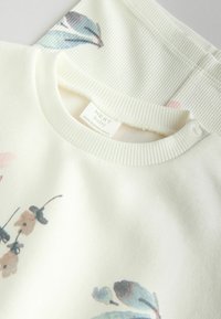 Creme sweatshirt med blomsterprint i bløde farver, ribbet krave med trykknaplukning og mærket "NEXT baby." Glat tekstur, afslappet design.