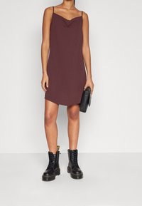 Lila glidklänning med tunna axelremmar och draperad nacke, matchad med svarta snörade ankelskor och en liten svart handväska.