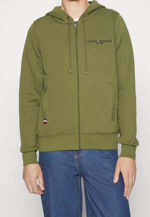 Mand iført en olivengrøn Tommy Hilfiger lynlåshoodie med logo på bryst og ærme, kombineret med blå denimjeans.