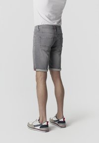 Mand iført grå denimshorts med opklebede kanter, hvid T-shirt og hvide, sorte og røde sneakers, stående med ryggen til på en ensfarvet baggrund.