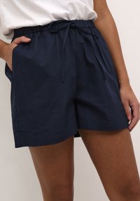 Pantaloni scurți bleumarin dintr-un material ușor, cu un elastic la talie și șnur, și buzunare laterale. Textură netedă, design casual.