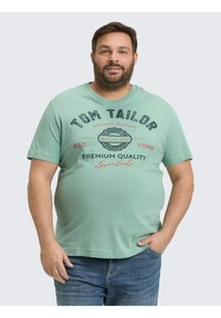 Mintgrünes T-Shirt mit kurzen Ärmeln, das mit gedrucktem Text in Marineblau und Rot versehen ist. Rundhalsausschnitt, weicher Stoff und bequemer Schnitt. Getragen mit blauen Jeans.